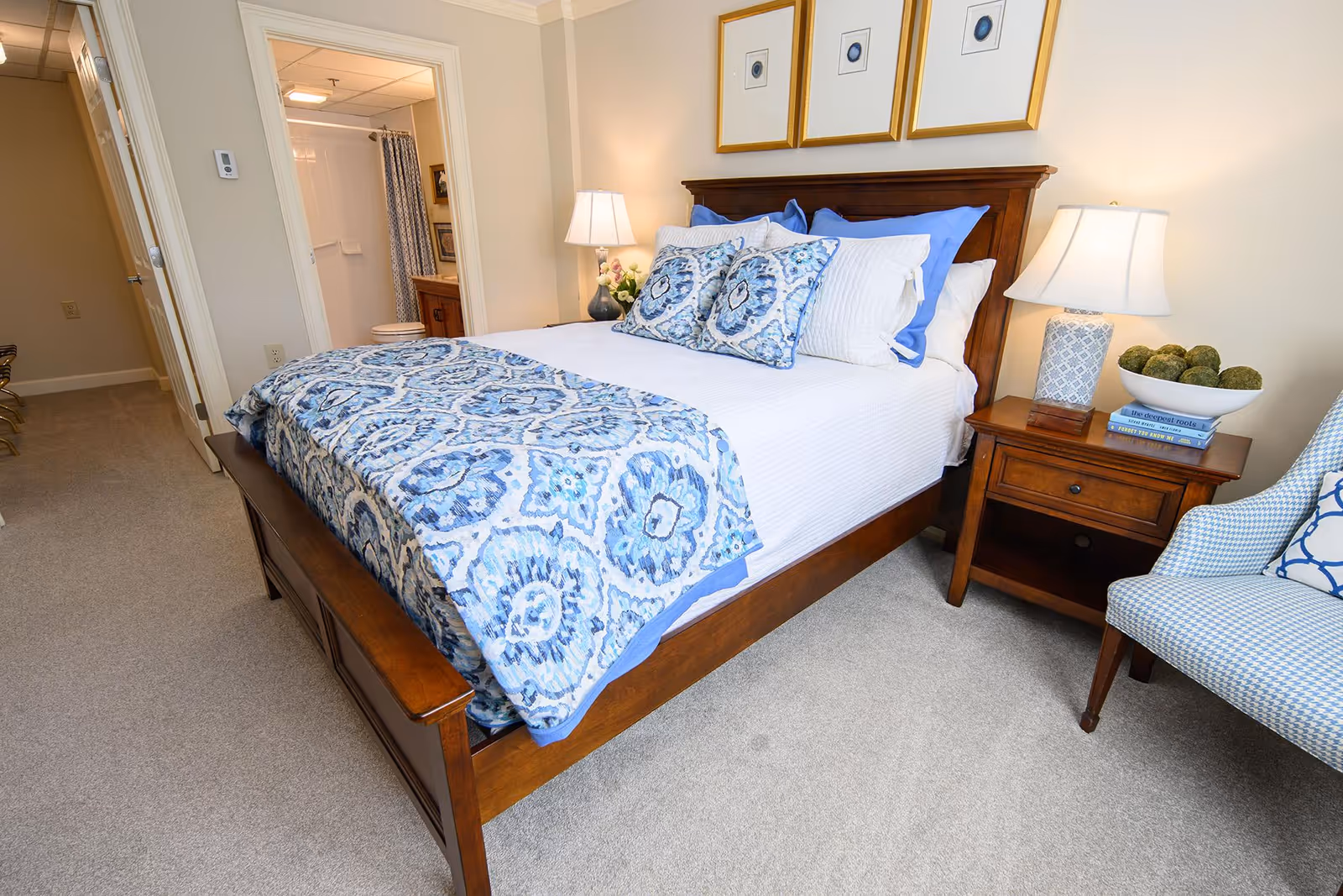 A neatly made bed with a wooden frame and blue patterned bedding in a bedroom. There are three framed pictures above the headboard. A wooden nightstand with a lamp, books, and decorative items is beside the bed. A blue patterned armchair is partially visible to the right. In the background, an open door reveals a bathroom with a shower curtain and vanity.