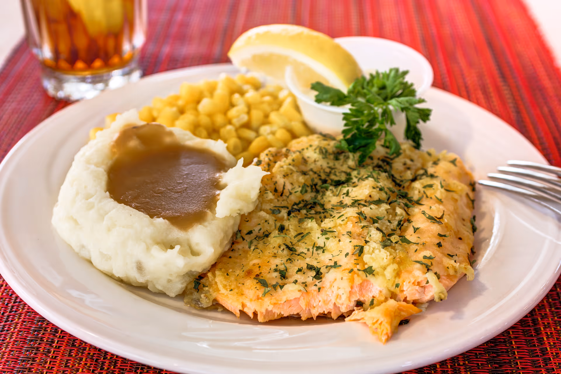 A plate of food featuring a serving of baked herb-crusted salmon, mashed potatoes with brown gravy, corn, a lemon wedge, and a sprig of parsley, placed on a red woven placemat with a glass of iced tea in the background.