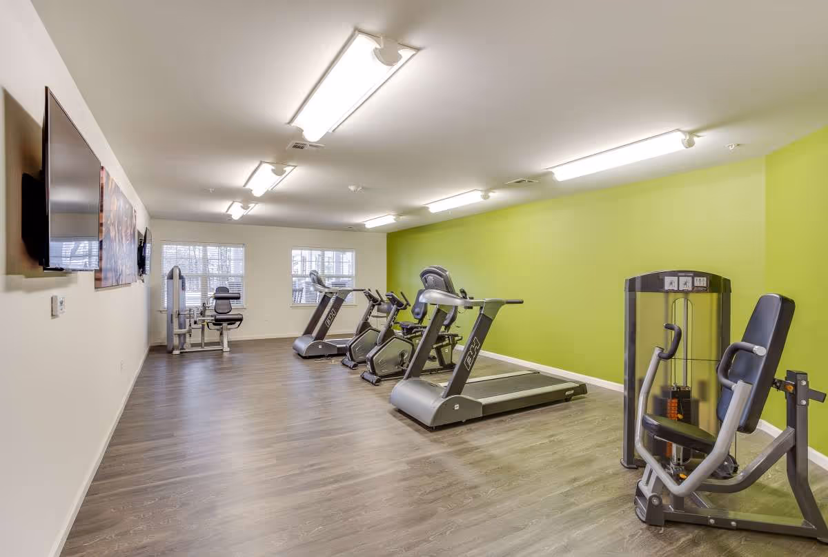 A fitness room with exercise equipment including treadmills, stationary bikes, and a weight machine. The room has a green accent wall, wood-style flooring, and bright overhead lighting. Two windows are visible at the far end, and a large TV is mounted on the opposite wall.