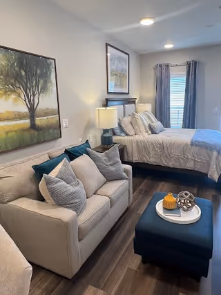 A furnished bedroom featuring a bed, sofa, ottoman with decor, bedside lamps and framed artwork by a window.