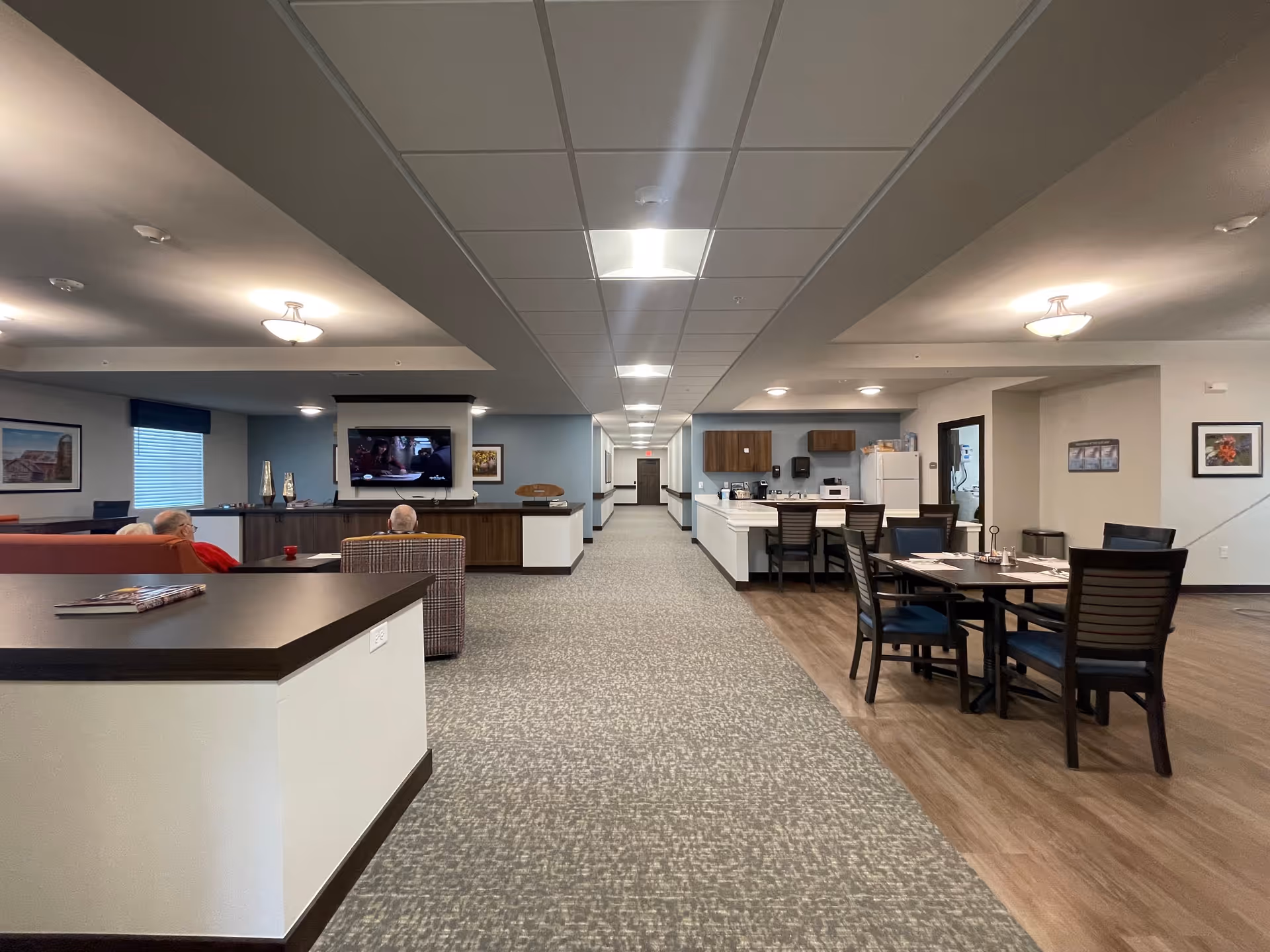 Open communal living area with seating and a wall-mounted TV on the left, dining tables and a kitchenette on the right, and a long central corridor down the middle.