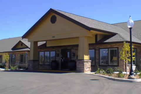 Front exterior view of a single-story building with a peaked roof and stone accents around the entrance. There are large windows on either side of the entrance and a street lamp on the right side. The building is surrounded by a paved driveway and some small landscaped plants.