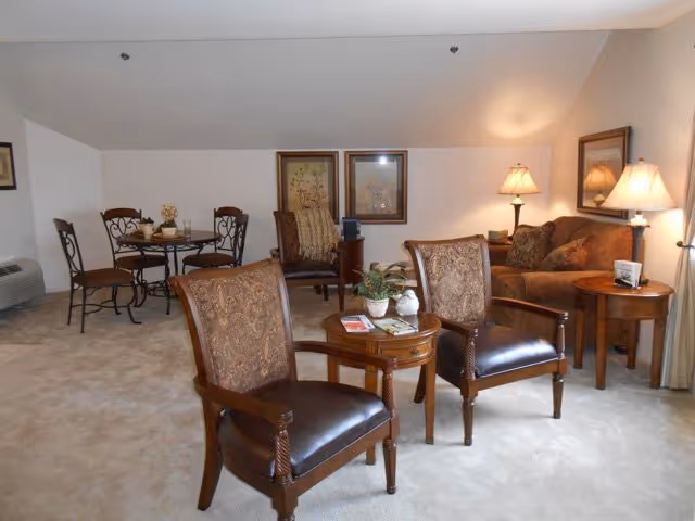 A cozy living room area with two upholstered armchairs and a small wooden table between them. In the background, there is a brown sofa with cushions, two table lamps on side tables, and framed artwork on the wall. To the left, a dining area with a round table and four chairs is visible. The room has beige carpeting and neutral-colored walls.