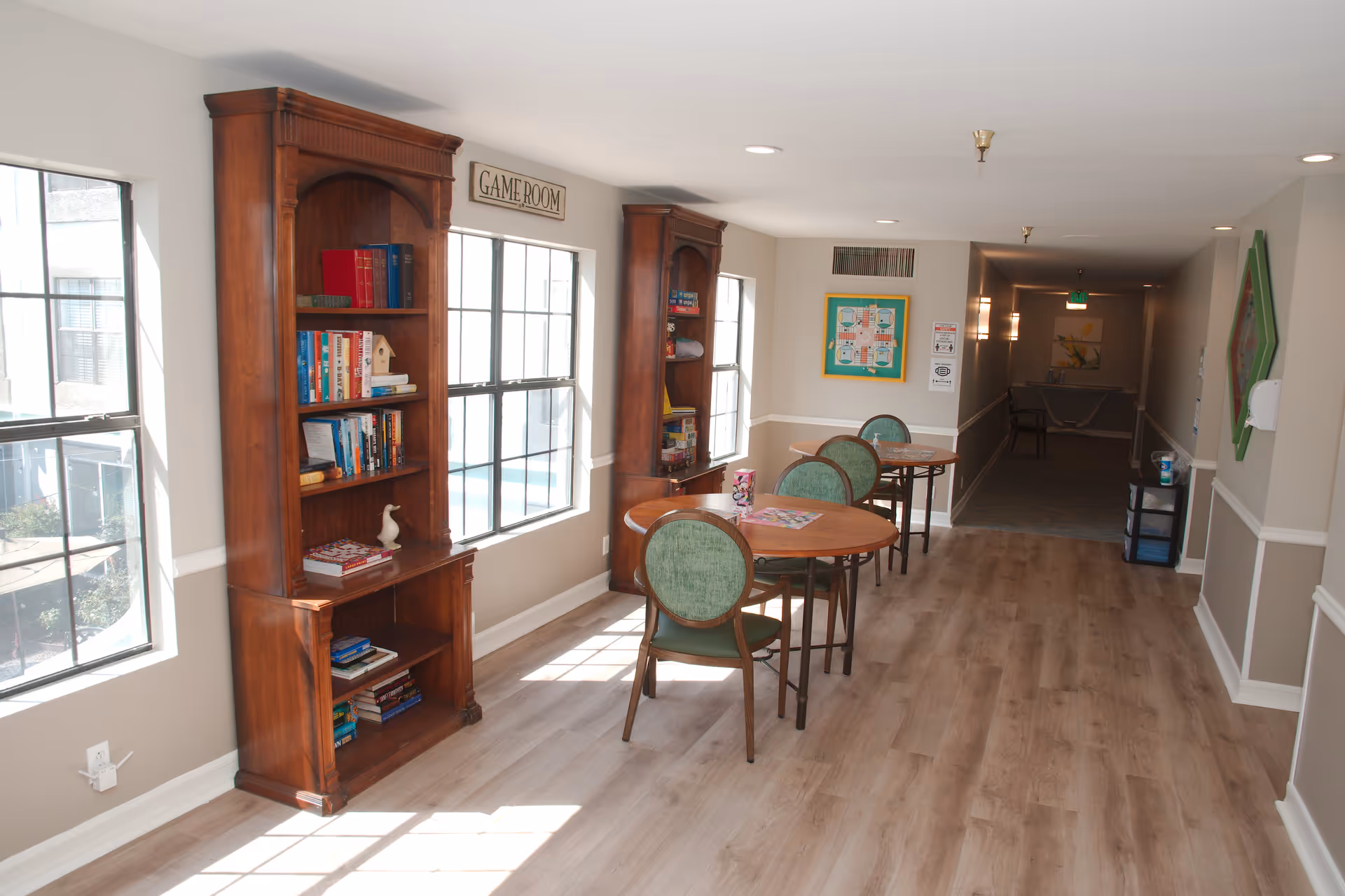 A bright hallway with large windows on the left side letting in natural light. Two wooden bookshelves filled with books and games are positioned against the wall beneath a sign that reads 'GAME ROOM'. Three round tables with green cushioned chairs are arranged along the hallway, each table having a game or puzzle on top. The floor is light wood, and the walls are painted beige with white trim. At the far end of the hallway, there is a corridor leading to another area with some wall decorations and an exit sign.