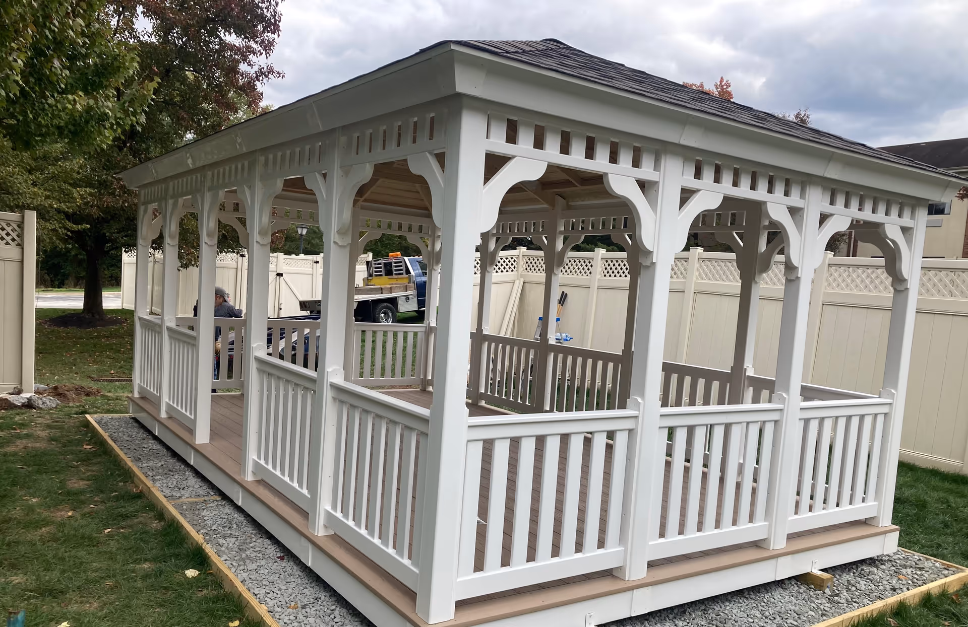 A white wooden gazebo with decorative railings and a shingled roof situated on a grassy area with a gravel border. There is a white privacy fence in the background and some trees with autumn foliage. A truck and a person are partially visible behind the gazebo.