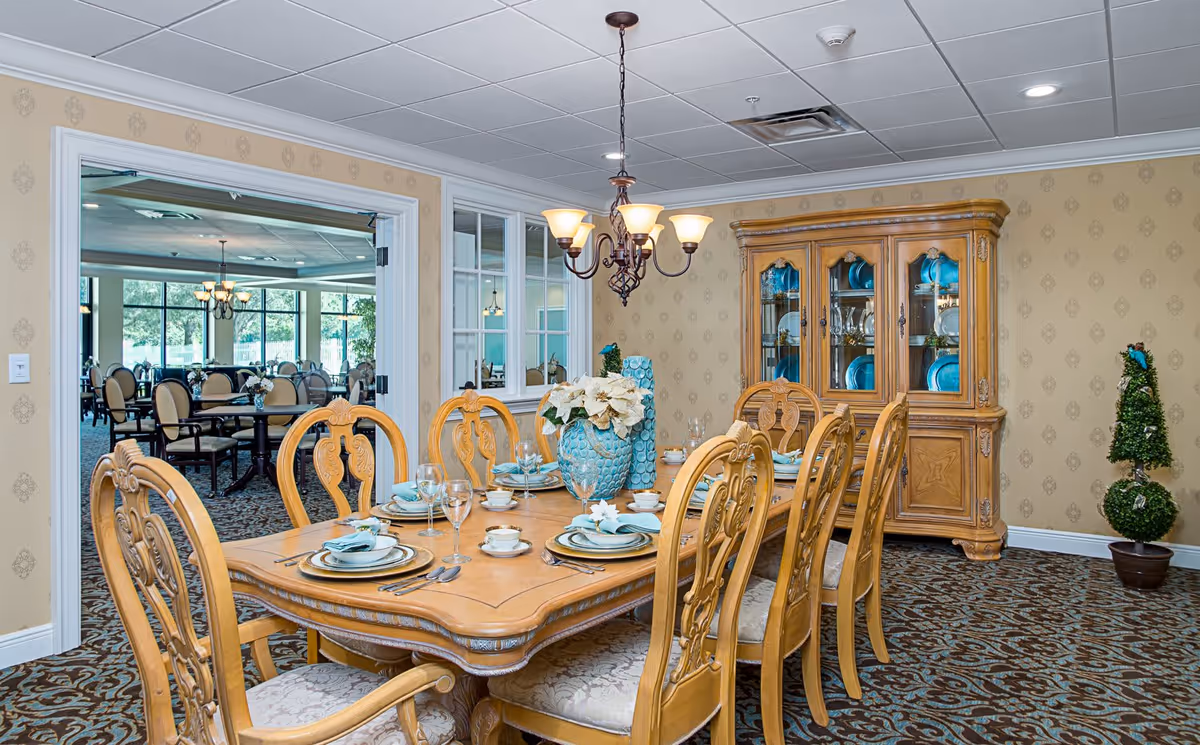 A formal dining room with a wooden dining table set for eight people. The table is decorated with blue and white dishware, glassware, and a centerpiece featuring white flowers in a blue vase. The room has patterned carpet, beige wallpaper with a subtle design, and a wooden china cabinet displaying blue plates and glassware. A chandelier with six lights hangs above the table. Through an open doorway, another dining area with multiple tables and chairs is visible, along with large windows showing greenery outside.