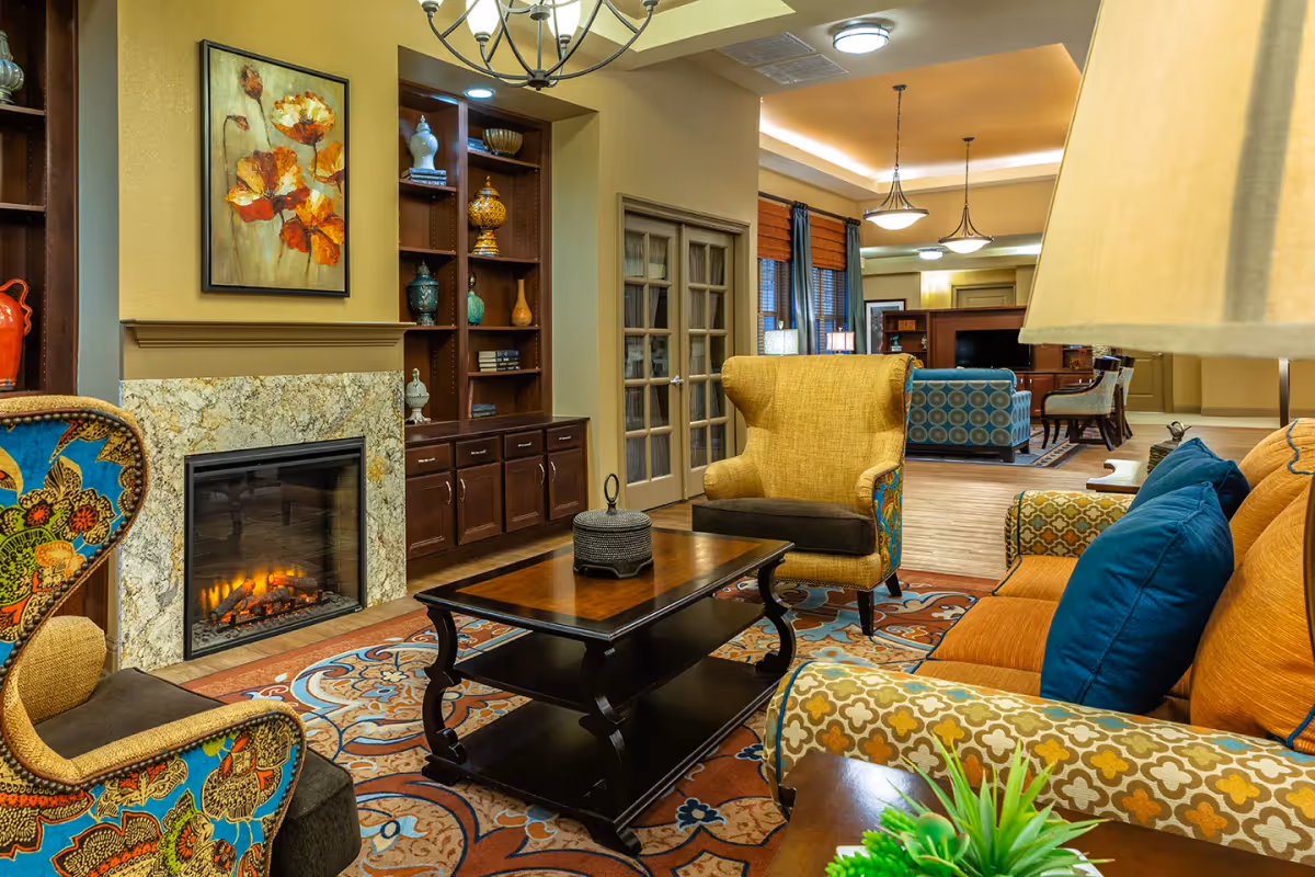 A cozy living room area in Sonoma House Assisted Living Alzheimer’s Care featuring a lit fireplace with a granite surround, colorful upholstered armchairs and sofa with blue cushions, a wooden coffee table on a patterned rug, built-in wooden shelves with decorative items, and warm lighting fixtures.