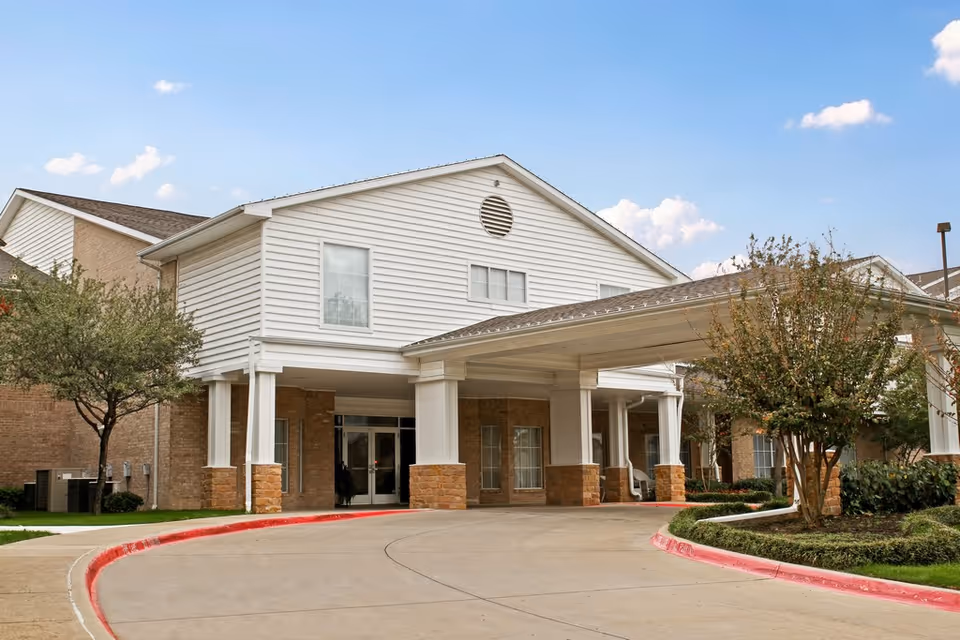 Exterior view of The Wellington at North Richland Hills senior living facility showing the entrance with a covered driveway, white siding, brick walls, and surrounding trees and landscaping under a blue sky with some clouds.