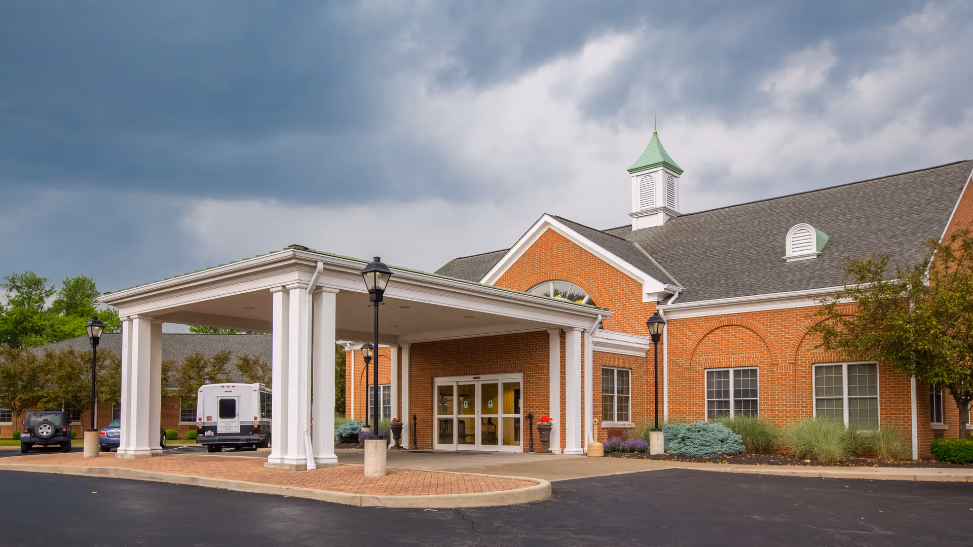 Front entrance of a red-brick senior living facility with a covered porte-cochere, lamp posts, parked vehicles, and a cloudy sky.