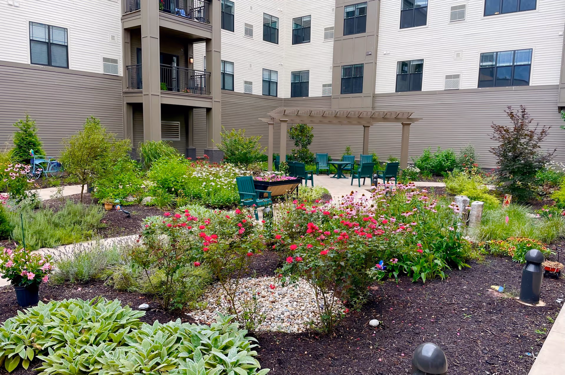 Landscaped courtyard with colorful flower beds, pathways, green chairs and a pergola in front of a multi-story residential building.