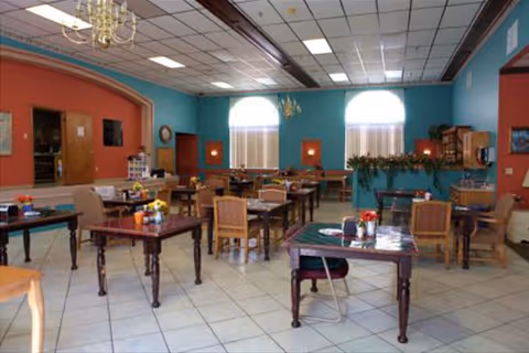 A spacious dining room with multiple wooden tables and chairs arranged neatly. The room features large windows with blinds, teal and coral colored walls, chandeliers hanging from the ceiling, and decorative plants on a shelf. The floor is tiled with light-colored tiles.