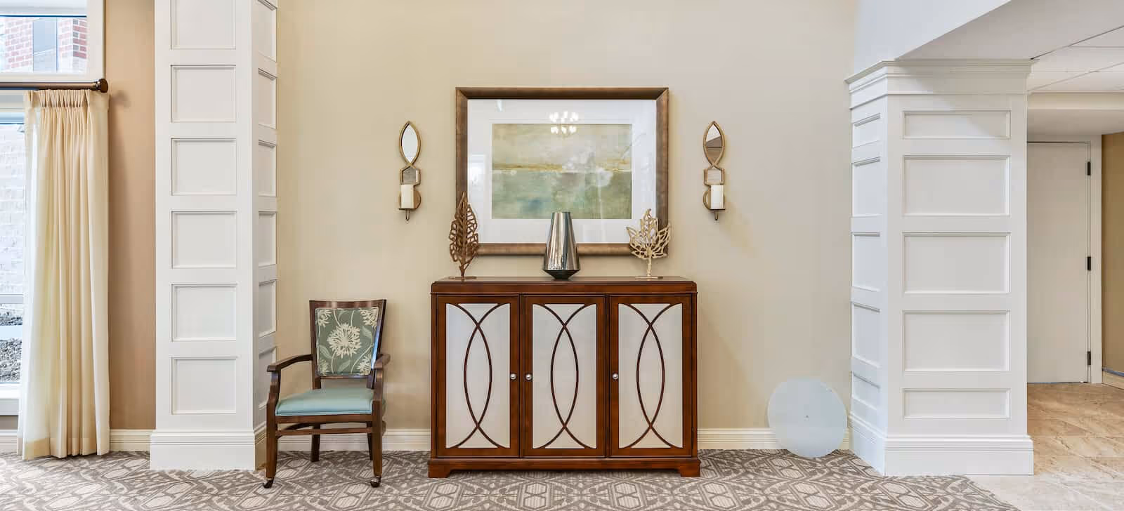 Bright interior common area with a wooden cabinet topped by artwork and decorative sconces, an upholstered chair, and white paneled columns.