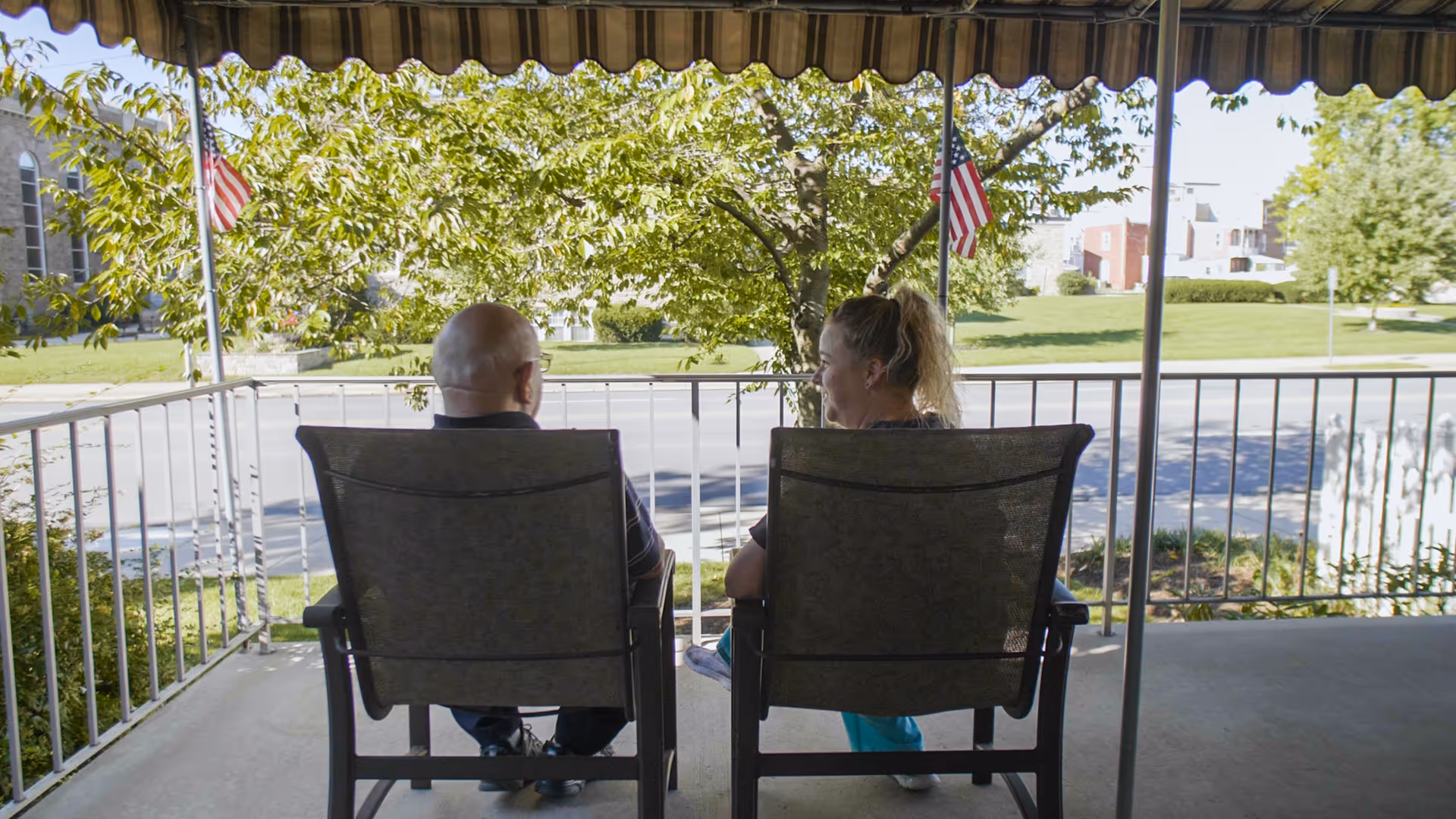 Two people sitting side-by-side in patio chairs on a covered porch overlooking a street and grassy lawn.