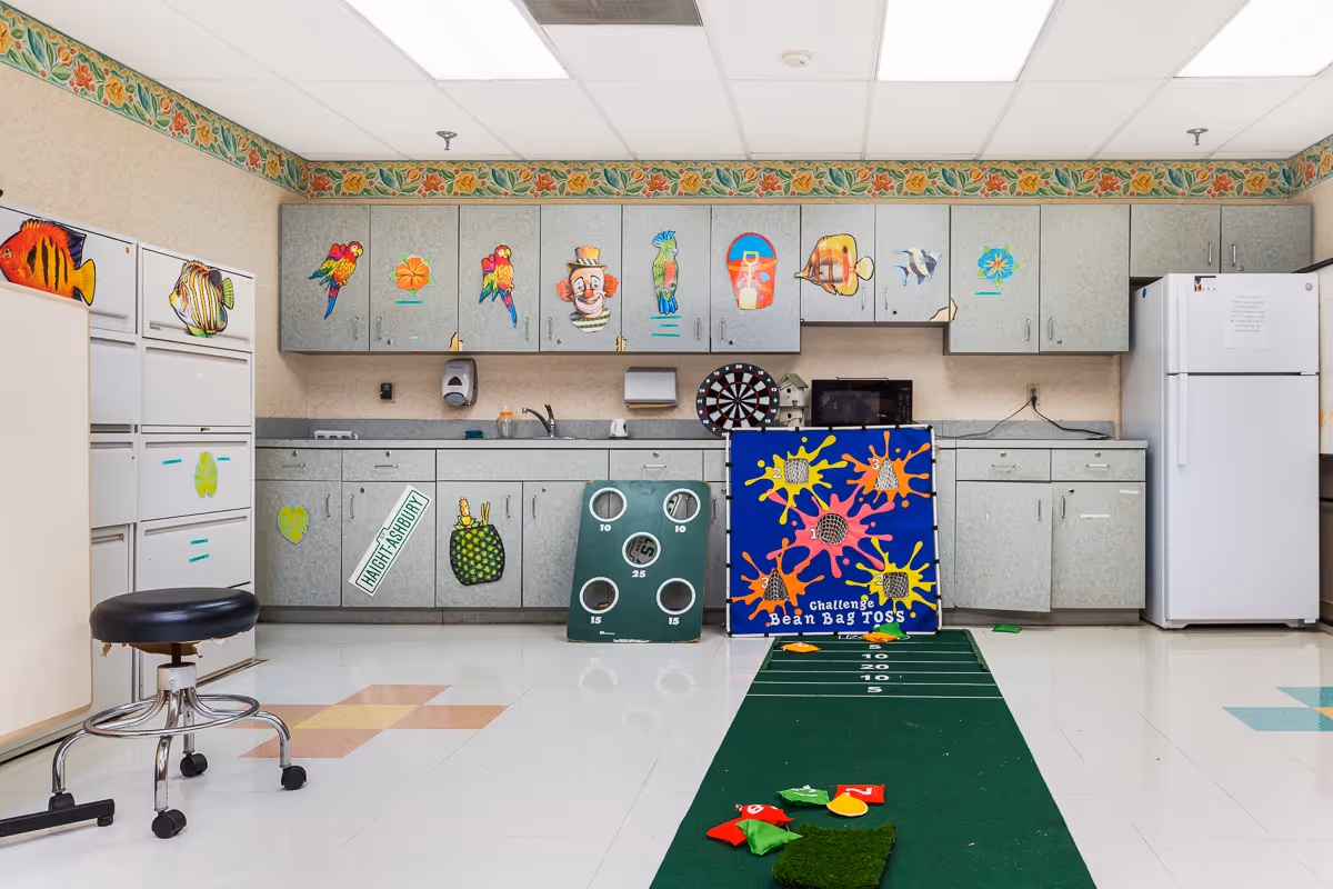 A kitchenette-style activity room with cabinets, a refrigerator, microwave and a bean bag toss game set out on a green mat.