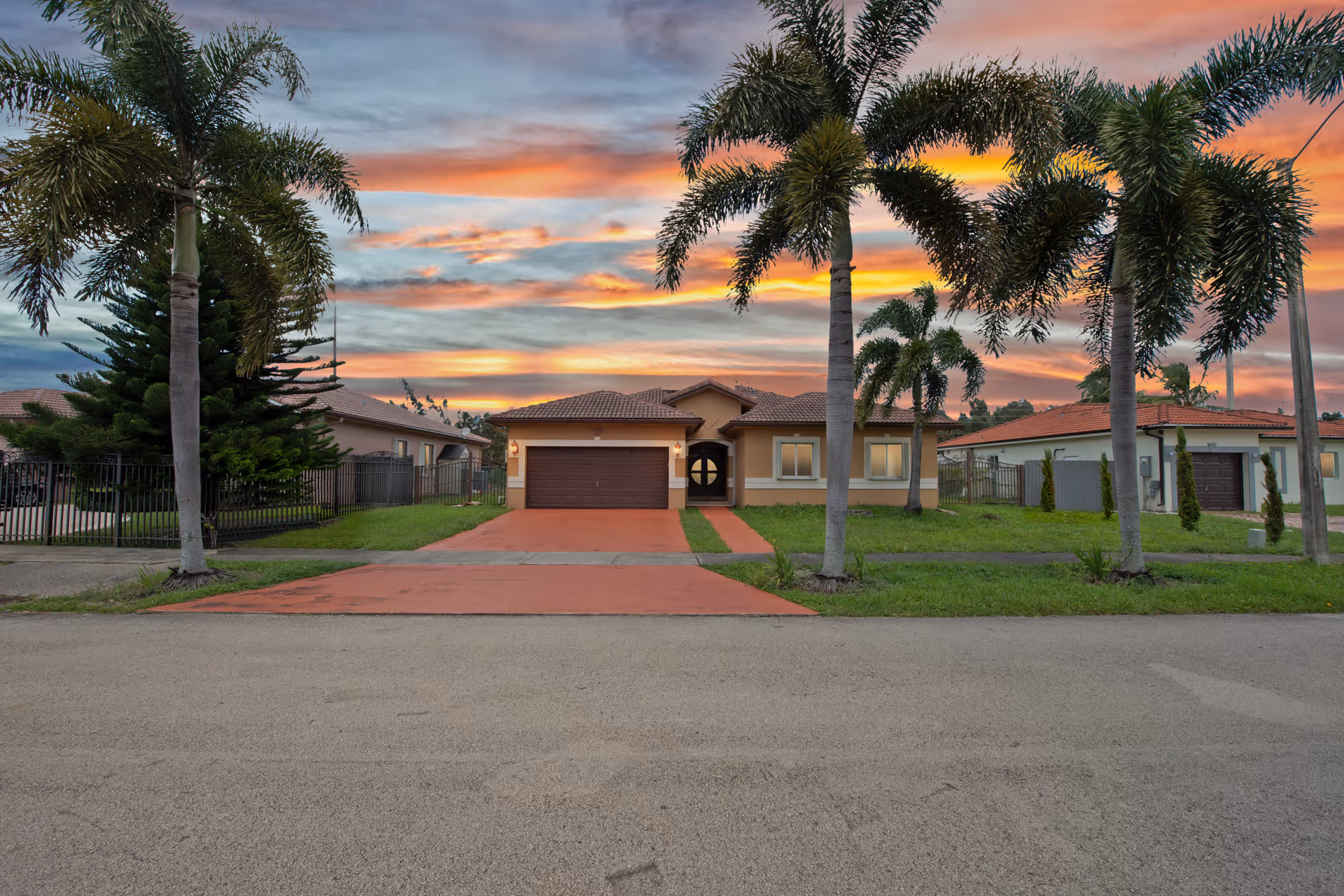 Front exterior view of a single-story residential building with a two-car garage, a red driveway, and palm trees in the front yard during a colorful sunset.