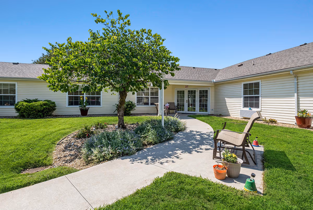 Outdoor courtyard area of Homestead Assisted Living of Emporia with a concrete pathway, green grass, a tree surrounded by plants, and a chair with potted plants nearby. The building exterior is light-colored with multiple windows and a double glass door entrance.