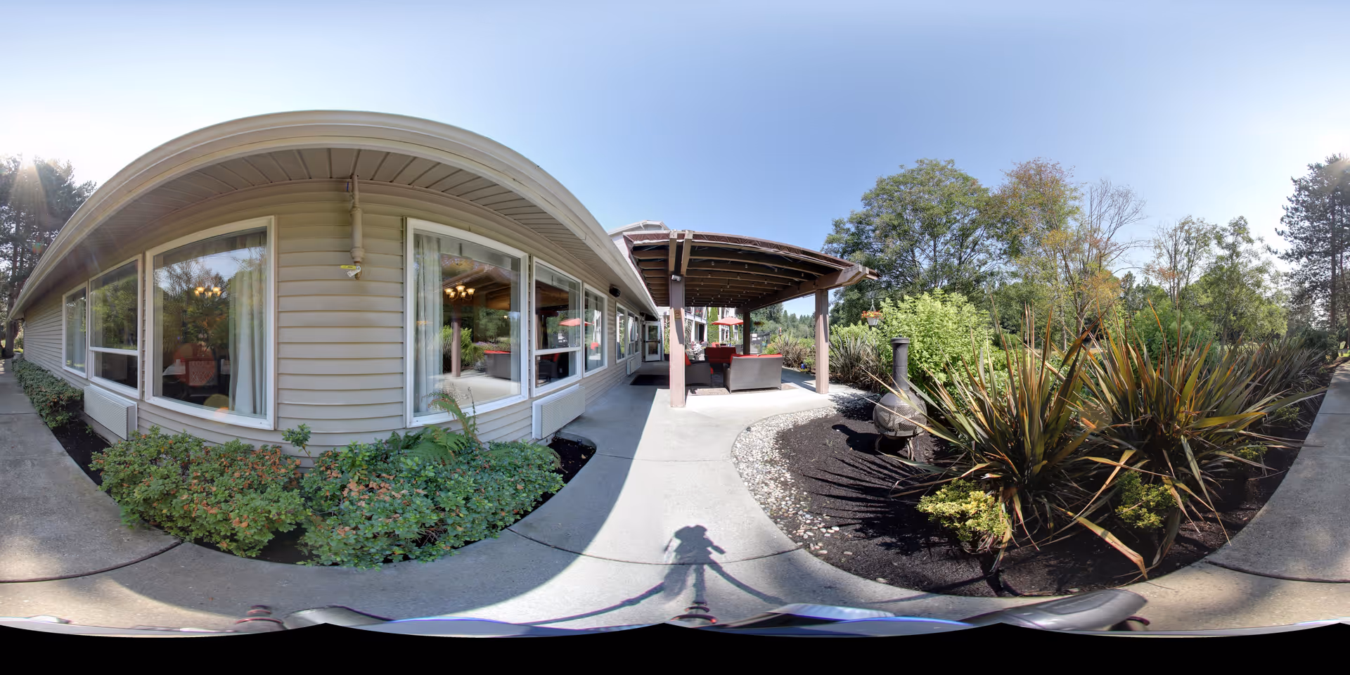 Outdoor patio area at Aegis Living Redmond featuring a covered seating area with chairs and a table, surrounded by landscaped garden beds with various shrubs and plants. The building exterior is visible with large windows and beige siding. The sky is clear and blue.