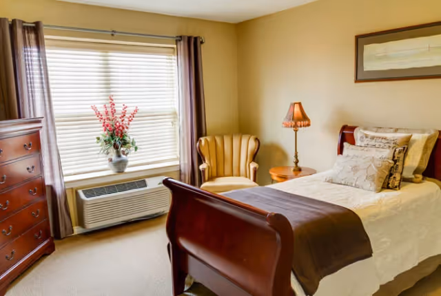 A cozy bedroom with a single wooden sleigh bed made with white bedding and decorative pillows. Next to the bed is a wooden nightstand with a vintage-style lamp. A comfortable upholstered armchair is placed in the corner near a window with blinds and brown curtains. A wooden dresser is positioned against the opposite wall, and a framed artwork hangs above the bed.
