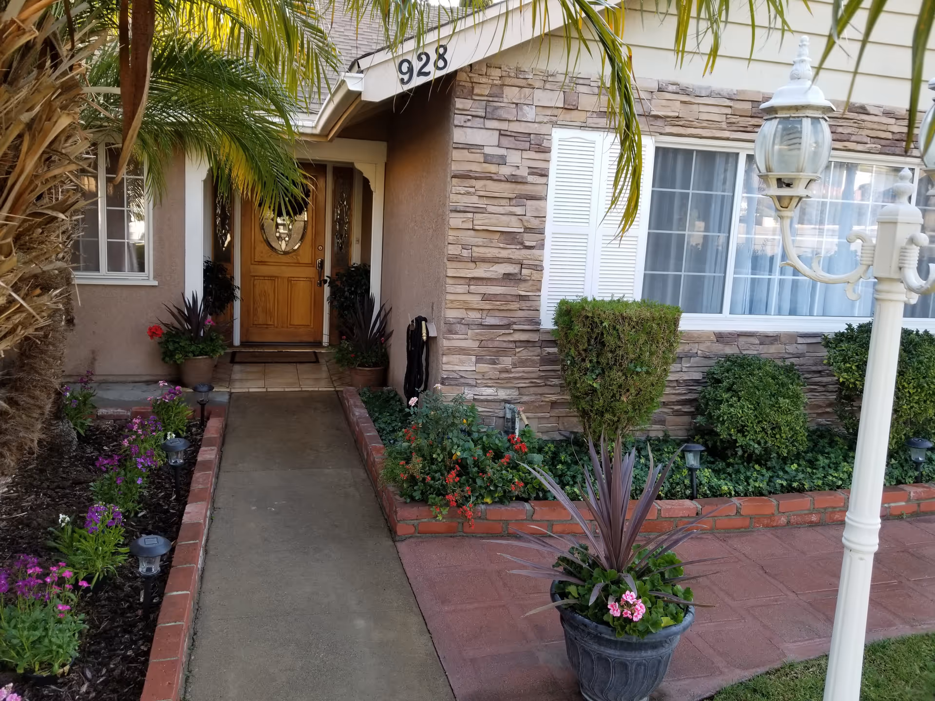 Front entrance of a house with a wooden door, surrounded by plants and flowers along a concrete pathway. The house exterior features stone and stucco walls, white window shutters, and a large window. A white lamp post is visible on the right side of the image.
