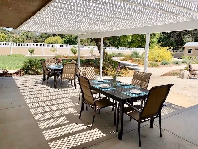 Outdoor patio area with two dining tables set with plates and glasses under a white lattice pergola casting patterned shadows on the ground. The patio overlooks a garden with green grass, shrubs, and trees, enclosed by a white fence.