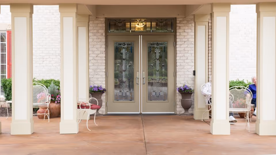 Covered entrance with double glass doors flanked by columns, potted flowers and outdoor chairs.