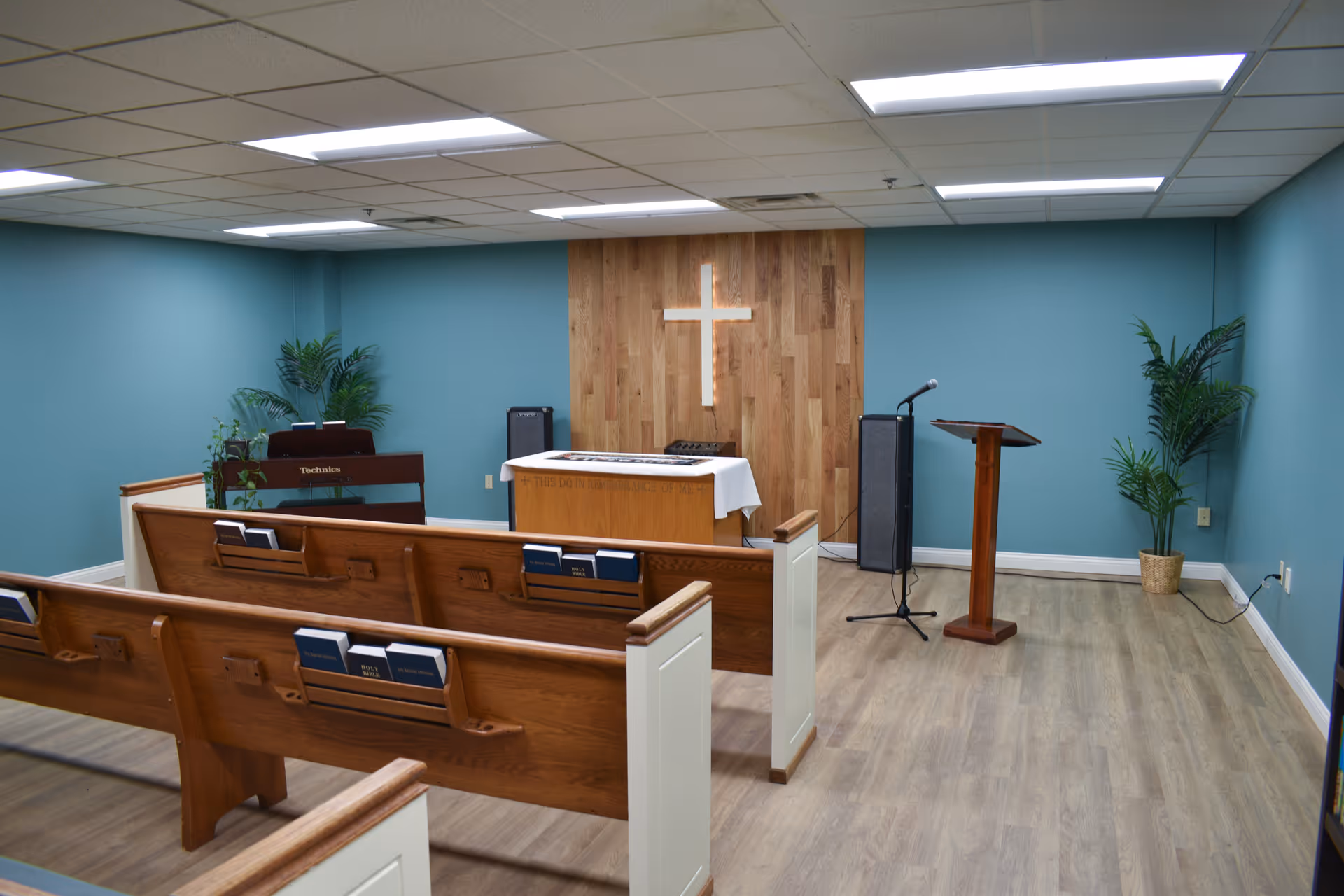 Interior of a small chapel or worship room with wooden pews, a wooden altar with a white cloth, a cross mounted on a wooden panel wall, a microphone on a stand, a lectern, a keyboard, and potted plants against teal-colored walls and a light wood floor.