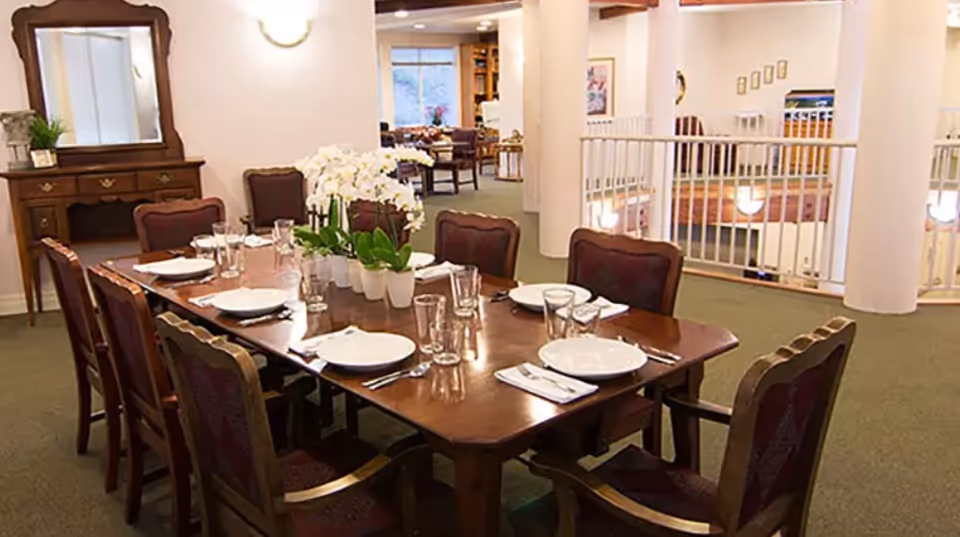 A formal dining table set for eight with plates, glasses, and a floral centerpiece in a communal senior living dining area.