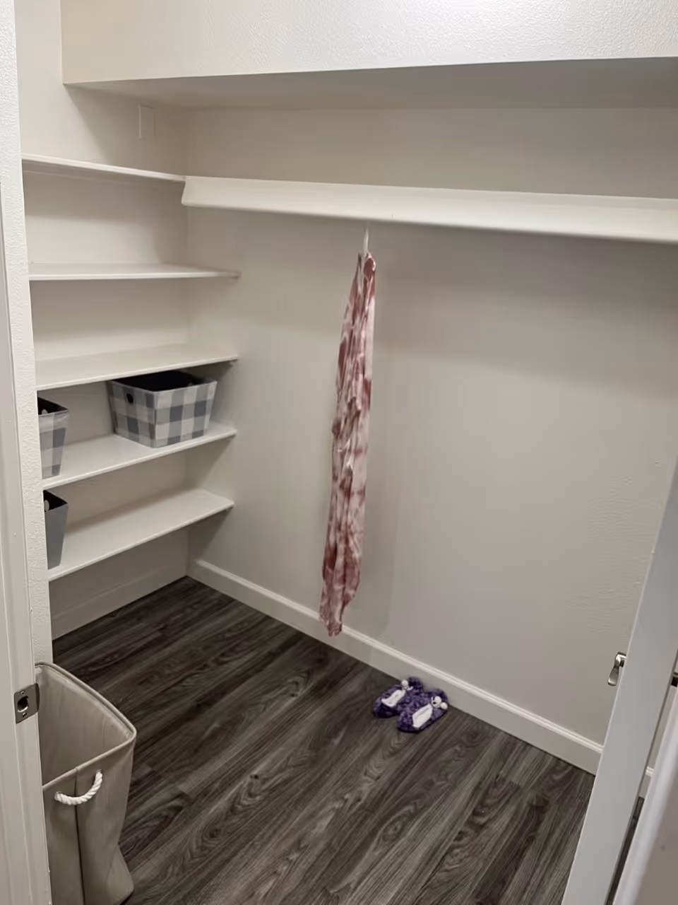 Empty walk-in closet with white walls and dark wood flooring. There are built-in white shelves on the left side holding three checkered storage bins. A pink and white tie-dye garment hangs from a rod on the right side, and a pair of purple slippers is on the floor near the back wall. A beige laundry hamper is partially visible in the foreground.