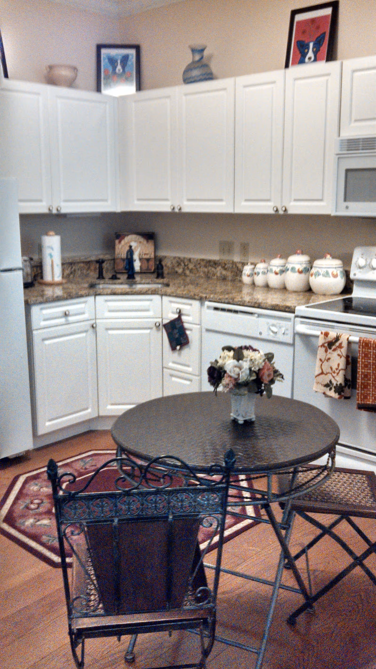 A cozy kitchen with white cabinets, granite countertops, and various kitchen items including a paper towel holder, decorative jars, and a stove with towels hanging on the handle. There is a round metal table with a floral centerpiece and two metal chairs with intricate designs. The floor has a wooden finish with a red patterned rug underneath the table.