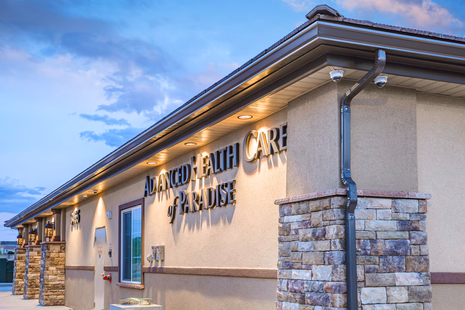 Exterior view of the Advanced Health Care of Paradise building during dusk, showing stone pillars, beige walls, a window, and the facility's name prominently displayed on the wall.