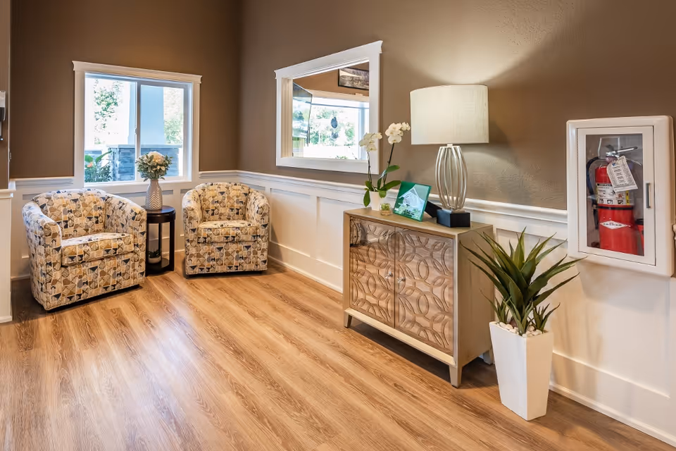 A cozy seating area with two patterned armchairs and a small round table with a flower vase between them, a wooden cabinet with decorative patterns, a table lamp, a potted plant, and a fire extinguisher mounted on the wall. The room has wood flooring, beige walls with white wainscoting, a window, and a large mirror reflecting the outside view.