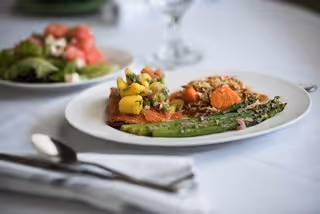 A close-up of a white plate with a meal consisting of grilled salmon topped with mango salsa, asparagus, and a serving of rice with carrots. In the background, there is a blurred plate with a salad and a glass of water on a white tablecloth.