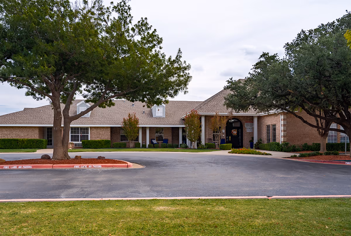 Single-story brick senior living facility entrance with driveway, trees, and manicured landscaping.