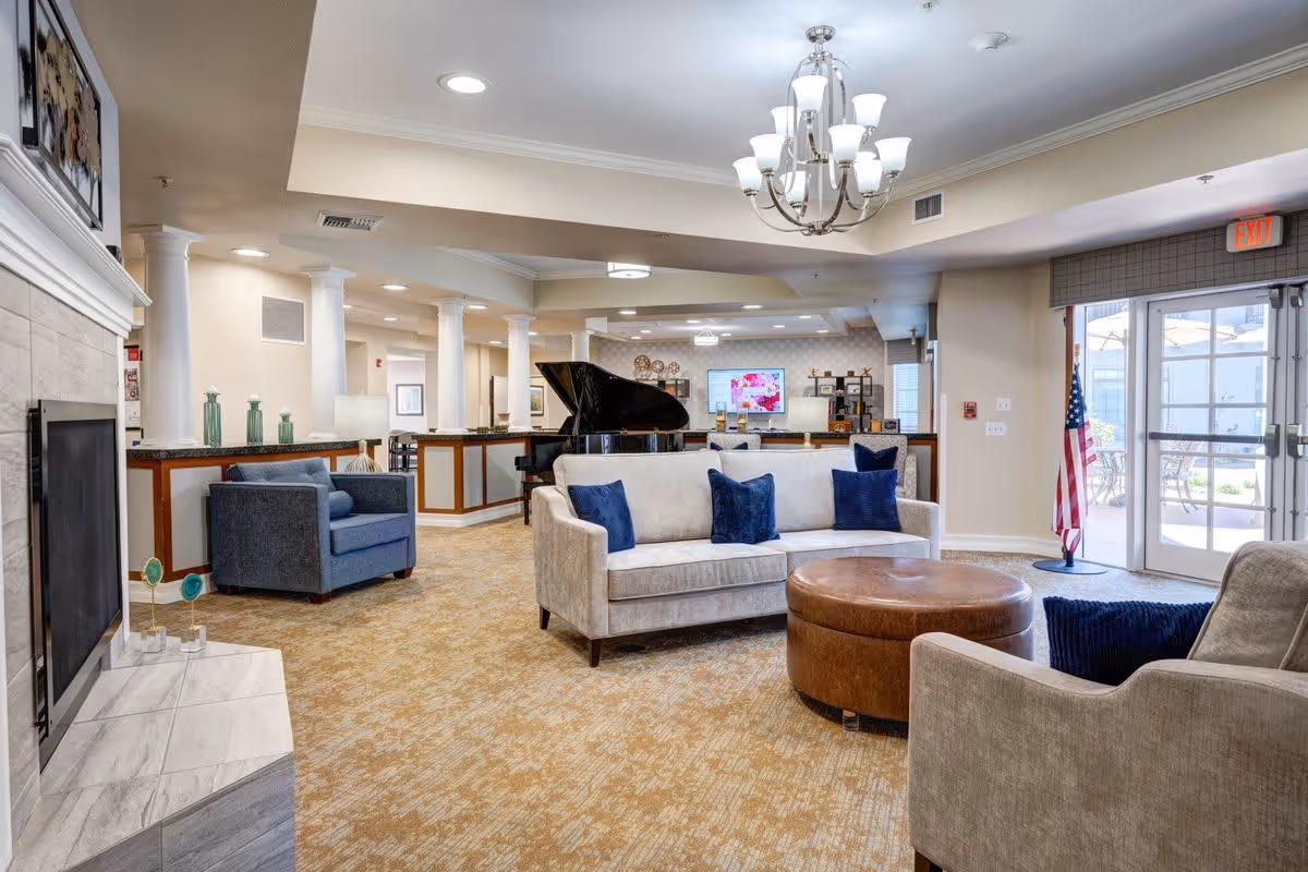 A spacious and well-lit living room area in a senior living facility featuring a beige sofa with blue cushions, a round brown ottoman, a blue armchair, a fireplace with a marble surround, a grand piano, and a chandelier. There are large windows and glass doors leading to an outdoor patio with tables and umbrellas. An American flag is positioned near the exit door.