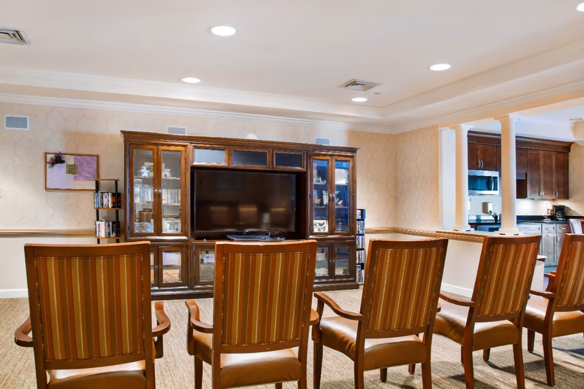 Rows of striped upholstered chairs facing a large TV and wooden entertainment cabinet in a well-lit common room adjacent to a kitchenette.