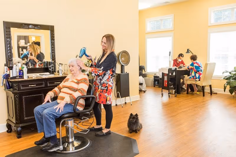 A senior woman sitting in a salon chair having her hair blow-dried by a stylist in a bright room with wooden floors. In the background, another woman is getting a manicure from a nail technician. A small black dog is sitting on the floor nearby.