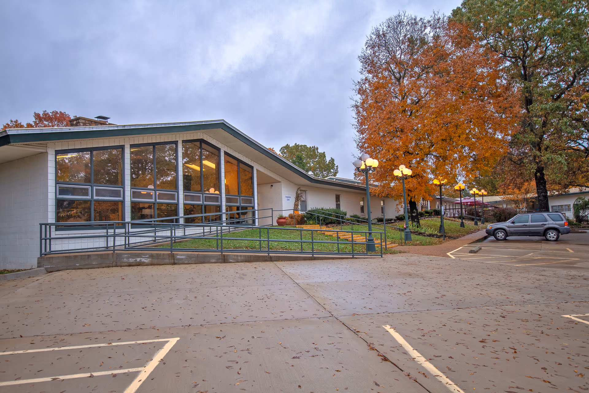 Single-story nursing center building front with large windows, an entrance ramp, parking area, and autumn trees.