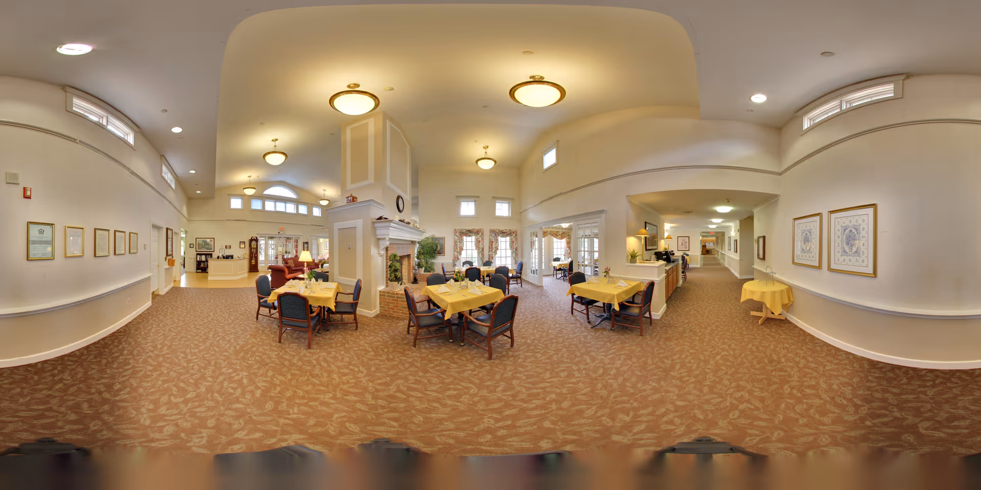 Spacious and well-lit common area in an assisted living facility featuring several tables with yellow tablecloths and chairs arranged around them. The room has high ceilings with multiple ceiling lights, large windows with curtains, a central fireplace, framed artwork on the walls, and a reception desk in the background. The carpet has a leaf pattern, and the overall atmosphere is warm and inviting.