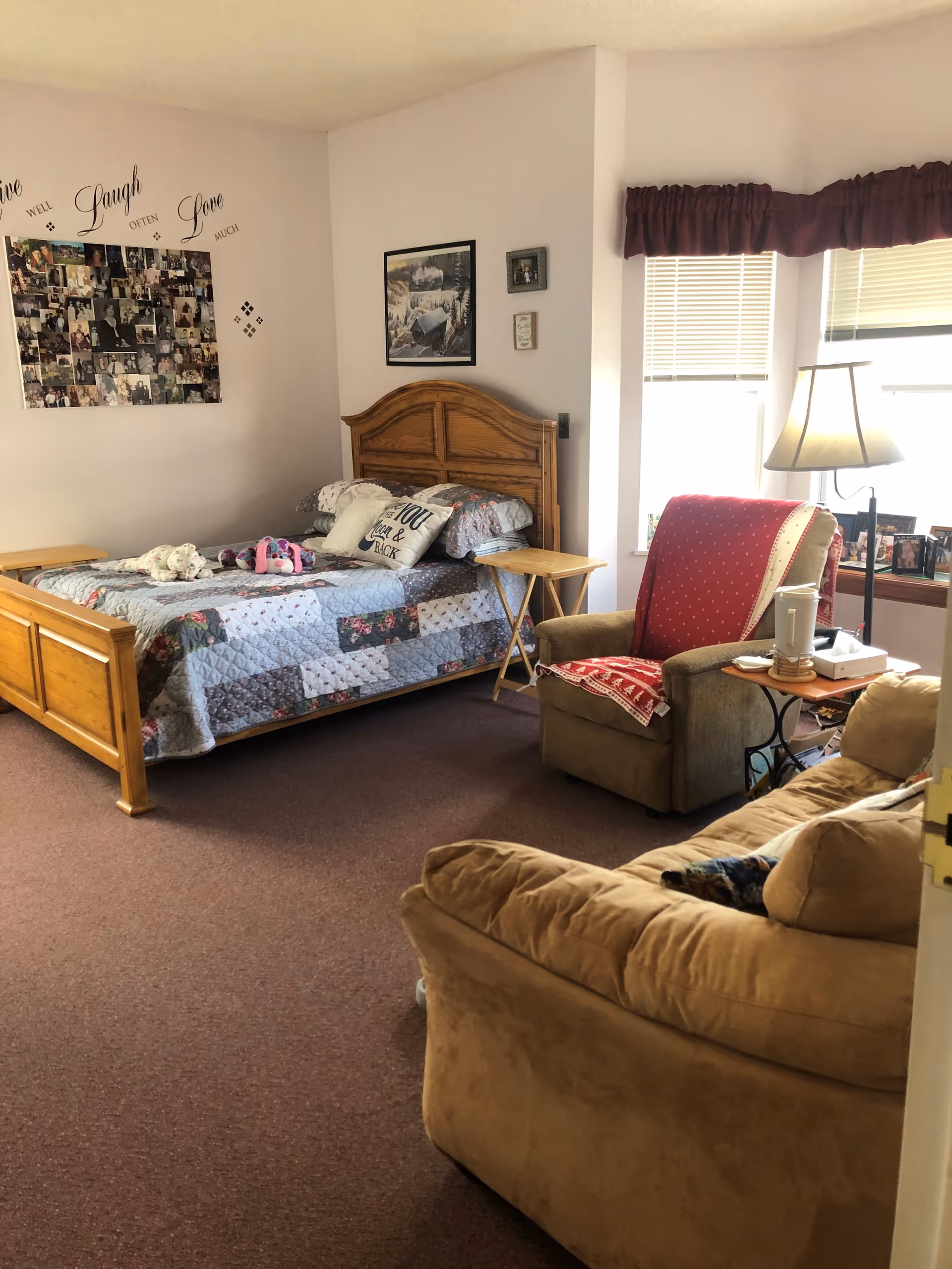 A cozy bedroom with a wooden bed frame, a patchwork quilt on the bed, and decorative pillows. There is a comfortable armchair with a red cover and a small side table with a lamp and some items on it. A beige couch is partially visible in the foreground. The walls are decorated with framed pictures and a collage of photos with the words 'Live Well Laugh Often Love Much' above it. Two windows with blinds and a maroon valance let in natural light.