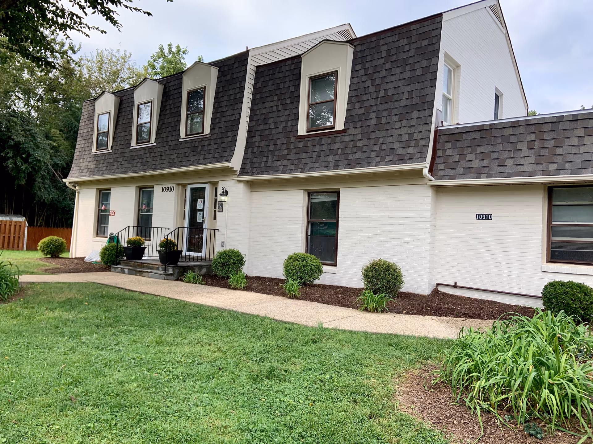 Exterior view of a two-story building with white brick walls and dark shingled mansard roof. The building has several windows and a small entrance with steps and black railings. There are green bushes and plants along the front, with a grassy lawn and a concrete walkway leading to the entrance.