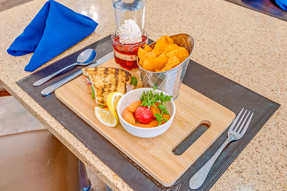A meal served on a wooden tray with a grilled sandwich, a small bowl of baby carrots and a radish garnished with parsley, a metal container of potato chips, a dessert cup topped with whipped cream and strawberries, and a glass of water. The tray is placed on a countertop with a blue folded napkin, a spoon, a knife, and a fork arranged around it.