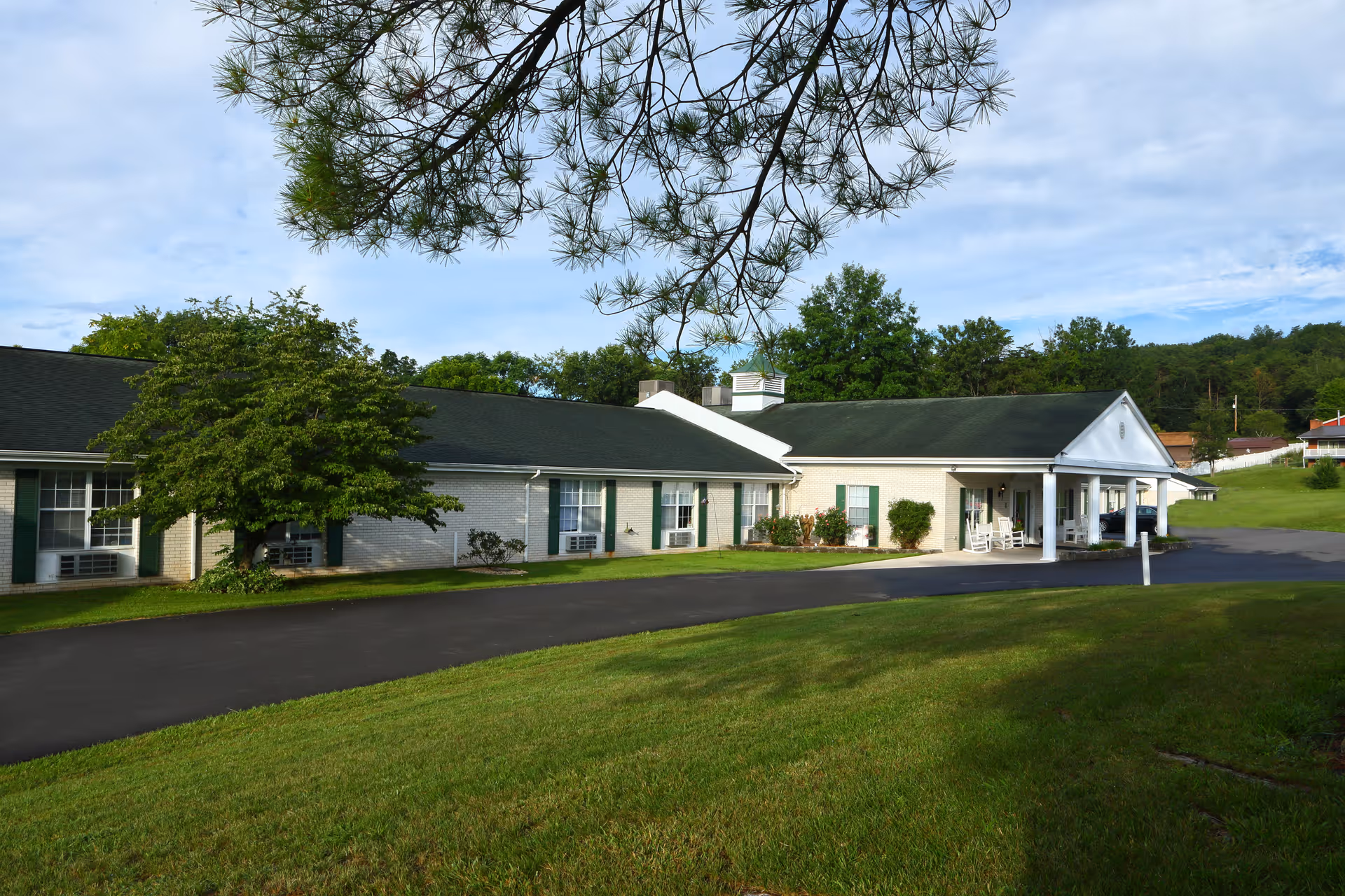 Single-story brick senior living building with a covered entrance, driveway and grassy lawn under a partly cloudy sky.