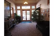 Interior view of a retirement facility lobby with a carpeted floor, a dark upholstered sofa on the right, a wooden cabinet on the left, several framed pictures on the walls, potted plants near the glass double doors leading outside, and a chandelier hanging from the ceiling.