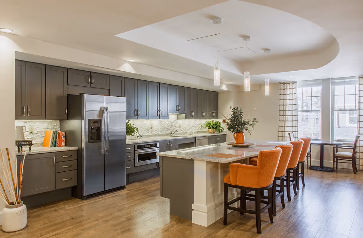 A modern kitchen with dark gray cabinets, a stainless steel refrigerator, built-in oven, and a large island with a granite countertop. Four orange upholstered chairs are placed at the island. The kitchen has wooden flooring and large windows with curtains letting in natural light. There are plants and decorative items on the countertops and island.