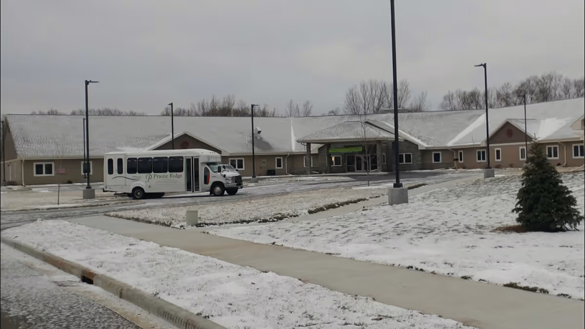 Exterior view of Prairie Ridge Assisted Living facility on a snowy day, showing a single-story building with a covered entrance and a white shuttle bus parked in front. There are street lamps and a sidewalk leading to the entrance, with snow covering the ground and rooftops.
