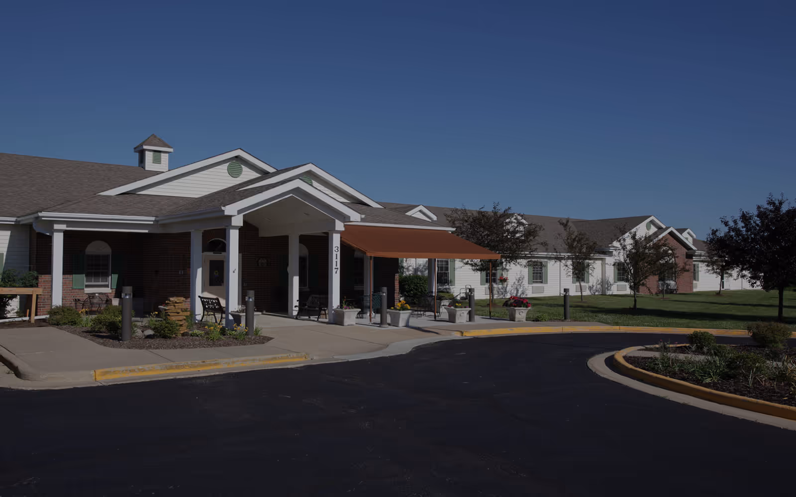 Front entrance of a single-story assisted living building with a covered porte-cochere, benches and a landscaped driveway.