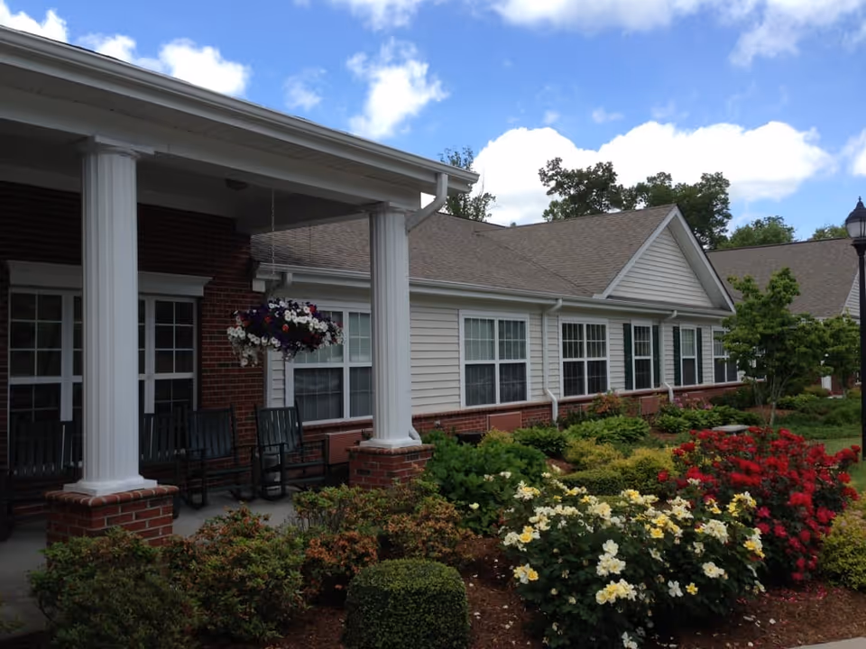 Front exterior of a single-story senior living building with a covered porch, white columns, rocking chairs and colorful landscaped flower beds.