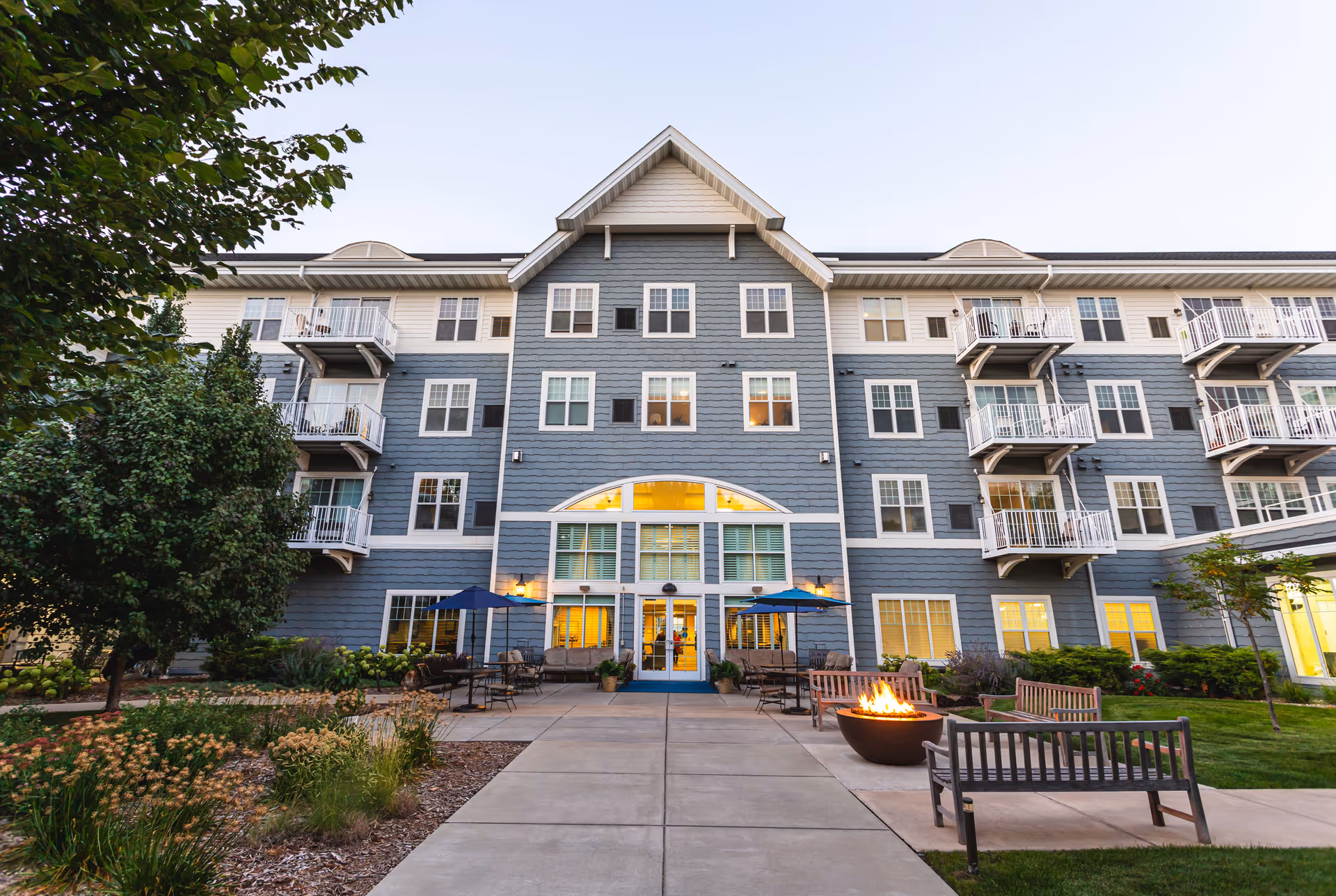 Front exterior of a multi-story senior living building with balconies, a central entrance flanked by lit windows, outdoor seating and a fire pit.