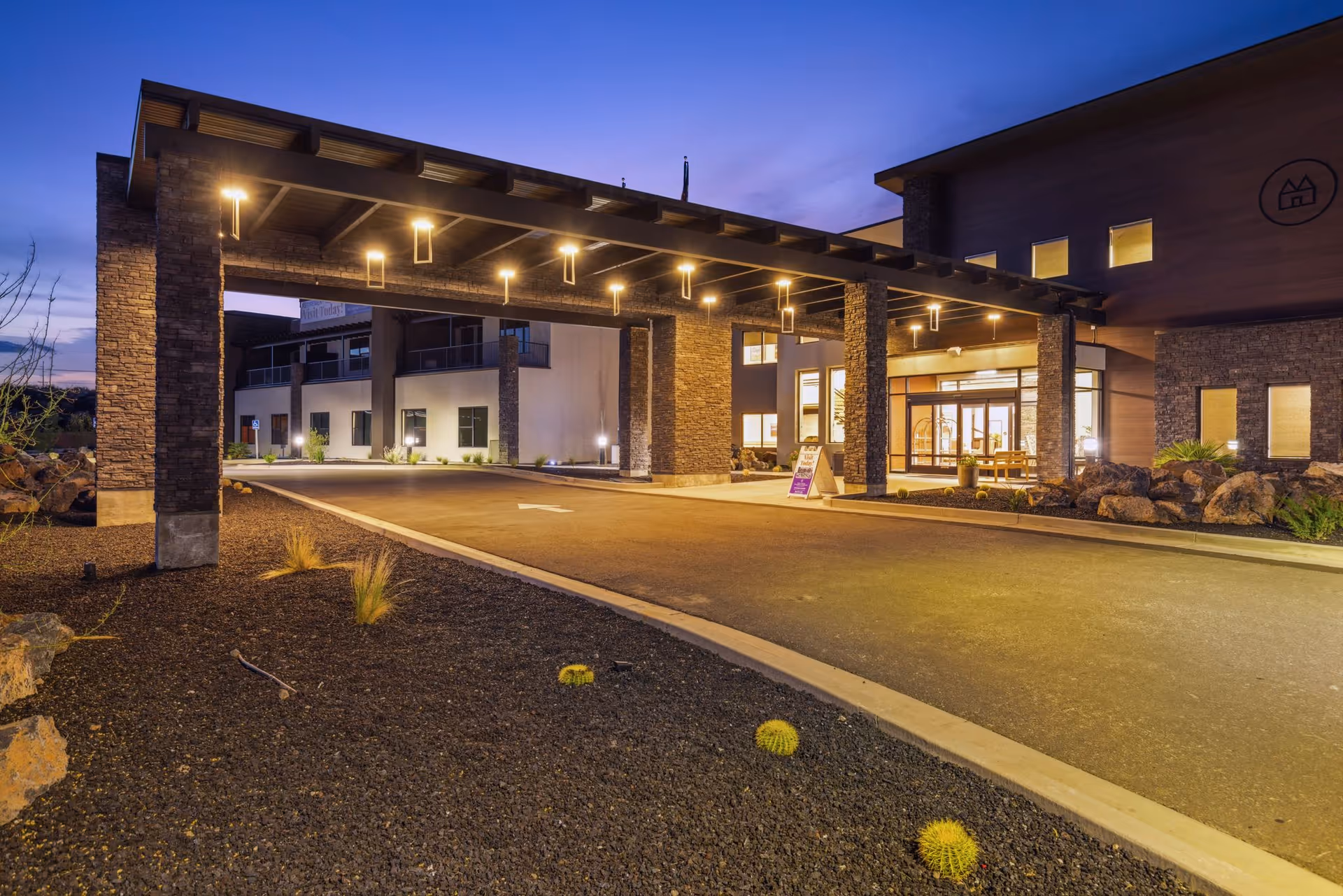 Illuminated entrance canopy and drop-off driveway of a retirement community building at dusk.