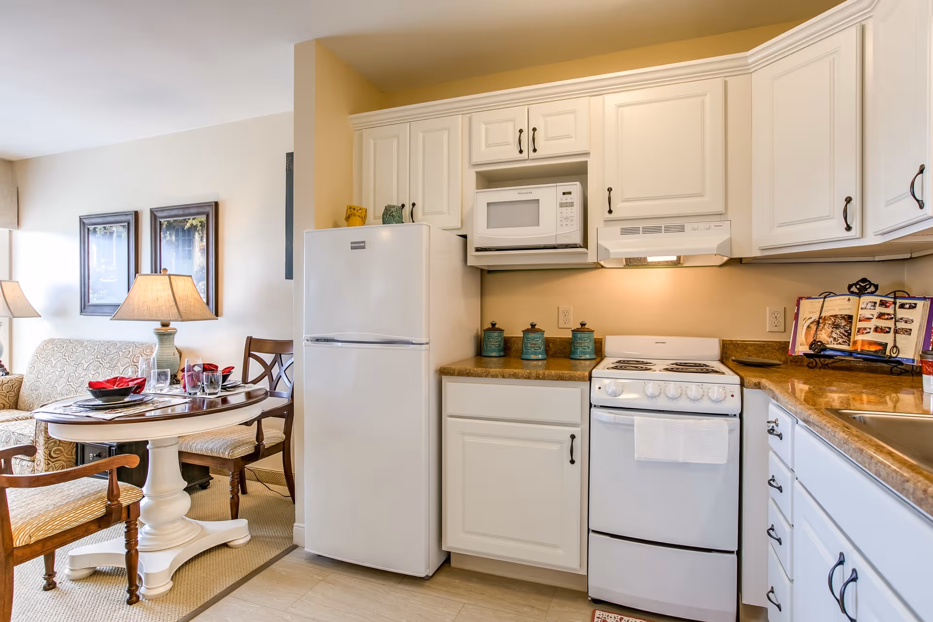 A bright and clean kitchen area with white cabinets, a white refrigerator, microwave, and stove. The kitchen counter has decorative jars and a cookbook on a stand. Adjacent to the kitchen is a small dining area with a round table set with plates, glasses, and red napkins, surrounded by wooden chairs and a cushioned sofa. Two framed pictures and a table lamp are visible in the background.